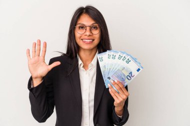 Beyaz arka planda izole edilmiş banknotları tutan genç Latin kadın gülümseyerek beş numarayı parmaklarıyla gösteriyor..