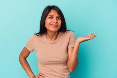 Genç Latin bir kadın, mavi arka planda izole edilmiş. Sorgulama sırasında omuz silkiyor ve şüphe ediyor..