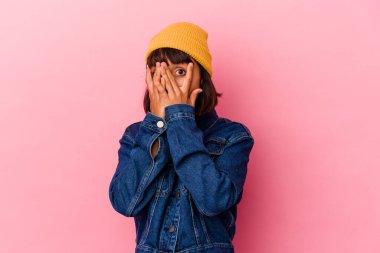 Pembe arka planda izole edilmiş genç melez kadın parmaklarının arasından korkmuş ve gergin bir şekilde göz kırpıyor..