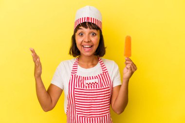 Genç melez dondurma üreticisi kadın sarı arka planda izole edilmiş dondurma tutuyor. Hoş bir sürpriz alıyor, heyecanlı ve el kaldırıyor..