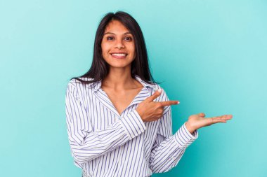 Mavi arka planda izole edilmiş genç Latin bir kadın, avucunun içinde bir fotokopi alanı tutarken heyecanlı..