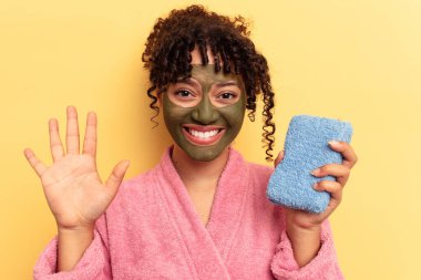 Sarı arka planda izole edilmiş duş süngeri tutan pembe bornozlu genç bir ırk kadını gülümseyerek beş numarayı parmaklarıyla gösteriyor..
