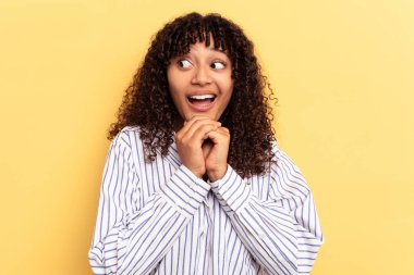 Genç melez kadın sarı arka planda izole edilmiş şans için dua ediyor, şaşırmış ve ağzı açık bir şekilde önüne bakıyor..