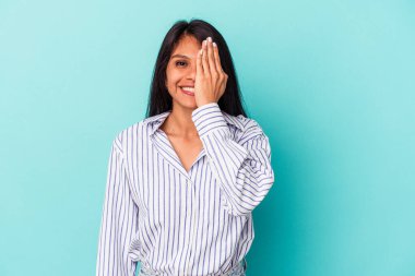 Mavi arka planda izole edilmiş genç Latin kadın yüzünün yarısını avuç içiyle kaplayarak eğleniyor..