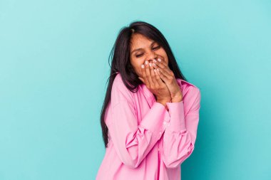 Mavi arka planda izole edilmiş genç Latin bir kadın bir şeye gülüyor, elleriyle ağzını kapatıyor..