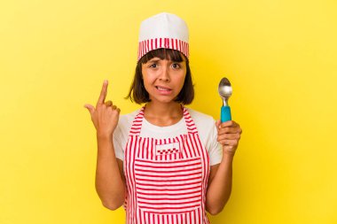 Sarı arka planda dondurmacı kadın dondurma tutuyor. Sarı arka planda izole edilmiş dondurma kepçesi tutan genç ırk dondurmacısı kadın işaret parmağıyla yaptığı hayal kırıklığı jestini gösteriyor..