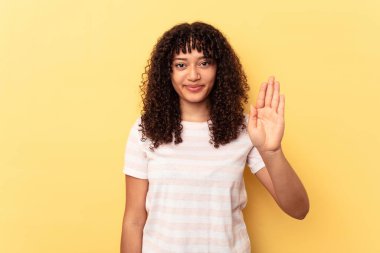 Sarı arka planda izole edilmiş genç bir ırk kadını gülümseyerek beş numarayı parmaklarıyla gösteriyor..