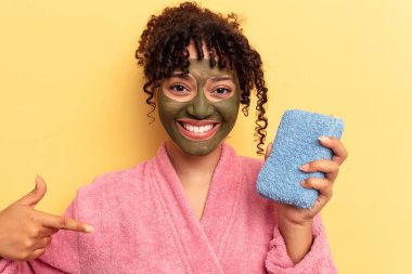 Pembe bornoz giyen genç bir ırk kadını sarı arka planda izole edilmiş bir duş süngeri tutuyor ve tişört fotokopi alanını elle gösteriyor, gururlu ve kendinden emin.