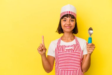 Sarı arka planda dondurmacı kadın dondurma tutuyor. Genç melez dondurma üreticisi kadın sarı arka planda izole edilmiş bir dondurma kepçesi tutuyor. Parmağıyla bir numarayı gösteriyor..