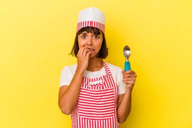 Genç melez dondurma üreticisi kadın elinde sarı arka planda izole edilmiş bir dondurma tutuyor. Genç melez dondurma üreticisi kadın sarı arka planda izole edilmiş, tırnak yiyen, gergin ve endişeli bir dondurma kepçesi tutuyor..