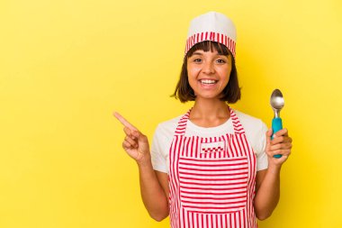 Genç melez dondurma üreticisi kadın elinde sarı arka planda izole edilmiş dondurma tutuyor. Küçük melez dondurma üreticisi kadın sarı arka planda izole edilmiş bir dondurma kepçesi tutuyor. Gülümsüyor ve boş alanda bir şey gösteriyor..