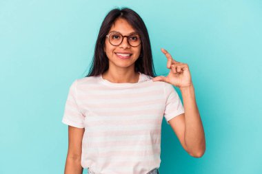 Mavi arka planda izole edilmiş genç Latin kadın işaret parmaklarıyla küçük bir şey tutuyor, gülümsüyor ve kendinden emin..