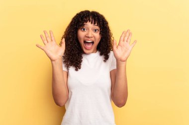Sarı arka planda izole edilmiş genç melez kadın hoş bir sürpriz yaşıyor, heyecanlı ve el kaldırıyor..