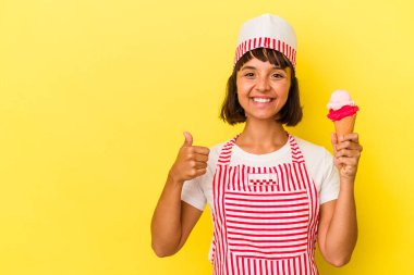 Genç melez dondurma üreticisi kadın dondurma tutuyor sarı arka planda izole edilmiş gülümsüyor ve başparmağını kaldırıyor.