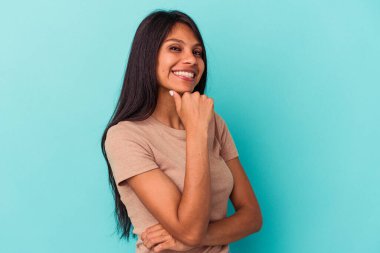Mavi arka planda izole edilmiş genç Latin kadın gülümsüyor ve kendinden emin, elleriyle çenesine dokunuyor..