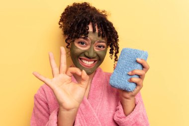 Pembe bornoz giyen, sarı arka planda izole edilmiş duş süngeri tutan, neşeli ve kendinden emin genç bir ırk kadını..