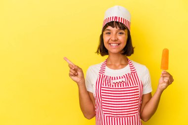 Sarı arka planda izole edilmiş dondurma tutan genç bir dondurma üreticisi kadın gülümsüyor ve boş alanda bir şey gösteriyor..
