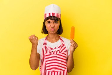 Küçük melez dondurma üreticisi kadın elinde dondurma tutuyor sarı arka planda izole edilmiş kameraya yumruğunu gösteriyor, agresif yüz ifadesi var..