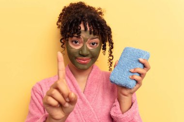 Pembe bornoz giyen, sarı arka planda izole edilmiş duş süngeri tutan genç bir ırk kadını parmaklarıyla bir numarayı gösteriyor..