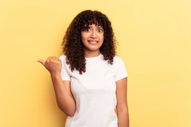 Sarı arka planda izole edilmiş genç melez kadın işaret parmağıyla fotokopi alanını işaret ederek şok oldu..