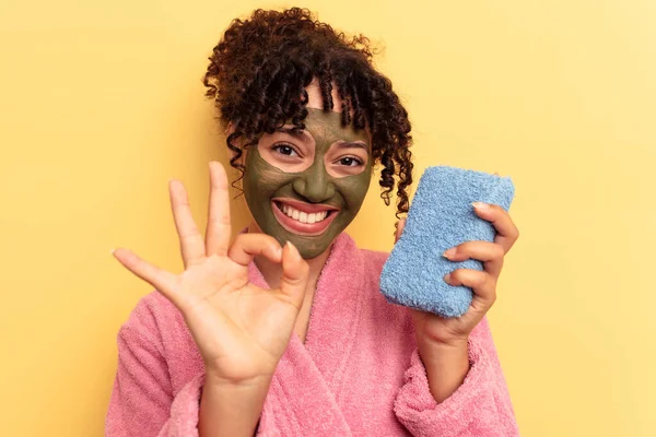 Pembe bornoz giyen, sarı arka planda izole edilmiş duş süngeri tutan, neşeli ve kendinden emin genç bir ırk kadını..