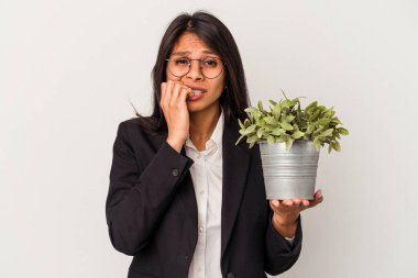 Beyaz arka planda bitkileri tutan genç Latin kadın tırnaklarını yiyor, gergin ve çok endişeli..