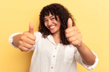 Sarı arka planda izole edilmiş genç bir ırk kadını. Baş parmağını kaldırıyor, gülümsüyor ve kendinden emin..