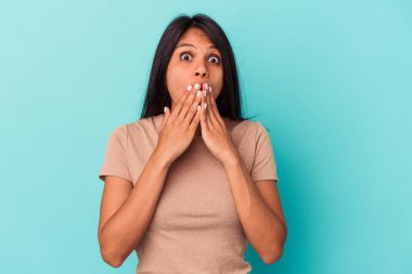 Mavi arka planda izole edilmiş genç Latin kadın elleriyle ağzını kapatırken şok oldu..