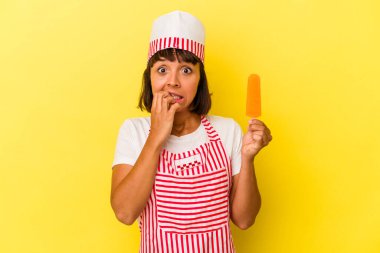 Genç melez dondurma üreticisi kadın elinde dondurma tutuyor sarı arka planda izole edilmiş tırnak yiyor, gergin ve endişeli..