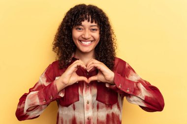 Sarı arka planda izole edilmiş genç melez kadın gülümsüyor ve elleriyle kalp şeklini gösteriyor..