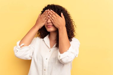 Sarı arka planda izole edilmiş genç melez kadın elleriyle gözlerini kapatmaktan korkuyor..