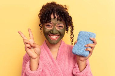 Pembe bornoz giyen, sarı arka planda izole edilmiş duş süngeri tutan genç bir ırk kadını iki numarayı parmaklarıyla gösteriyor..