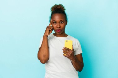 Elinde cep telefonu olan genç bir Afrikalı Amerikalı kadın mavi arka planda izole edilmiş bir şekilde tapınağı işaret ediyor, düşünüyor ve bir göreve odaklanıyor..