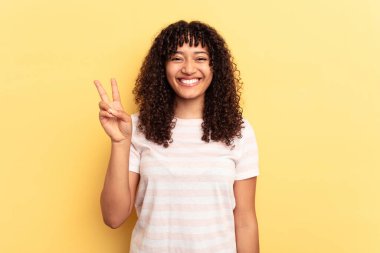 Sarı arka planda izole edilmiş genç melez kadın neşeli ve tasasız parmakları olan bir barış sembolü gösteriyor..