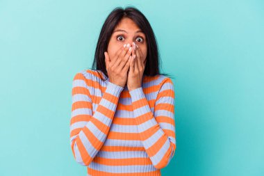 Mavi arka planda izole edilmiş genç Latin kadın şok olmuş, ağzı ellerle kaplanmış, yeni bir şey keşfetmek için sabırsızlanıyor..