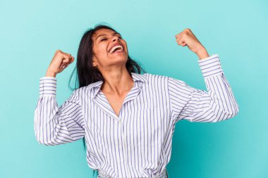 Genç Latin kadın mavi arka planda izole edilmiş zafer sonrası yumruğunu kaldırıyor, kazanan konsept..