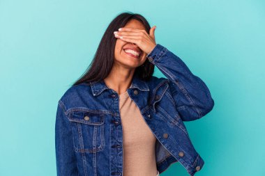 Mavi arka planda soyutlanmış genç Latin bir kadın elleriyle gözlerini kapatıyor, geniş bir gülümsemeyle bir sürpriz bekliyor..