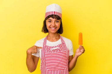 Genç melez dondurma üreticisi kadın, sarı arka plandaki izole edilmiş dondurmayı tutuyor tişört fotokopi alanını işaret ediyor, gururlu ve kendinden emin.