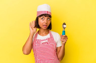 Genç melez dondurma üreticisi kadın, sarı arka planda izole edilmiş bir dondurma tutuyor. Küçük melez dondurma üreticisi kadın, sarı arka planda izole edilmiş bir dondurma kepçesiyle bir dedikoduyu dinlemeye çalışıyor..