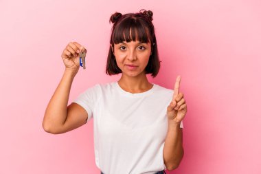 Genç melez bir kadın pembe arka planda izole edilmiş bir anahtar tutuyor. Parmağıyla bir numarayı gösteriyor..