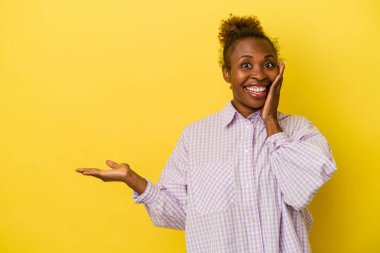Sarı arka planda izole edilmiş genç Afrikalı Amerikalı kadın avuç içinde fotokopi alanı tutuyor, el üstünde tutuyor. Şaşırmış ve mutlu..