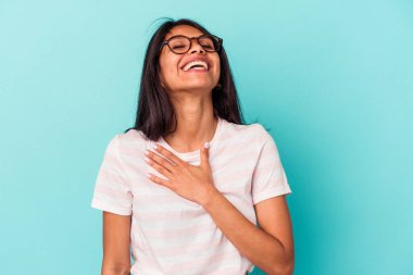 Mavi arka planda izole edilmiş genç Latin kadın yüksek sesle gülüyor elini göğsüne koyuyor..