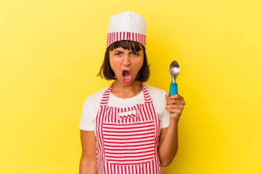 Genç melez dondurma üreticisi kadın elinde sarı arka planda izole edilmiş bir dondurma tutuyor. Genç melez dondurma üreticisi kadın sarı arka planda izole edilmiş bir dondurma kepçesi tutuyor. Kızgın ve agresif bir şekilde bağırıyor..