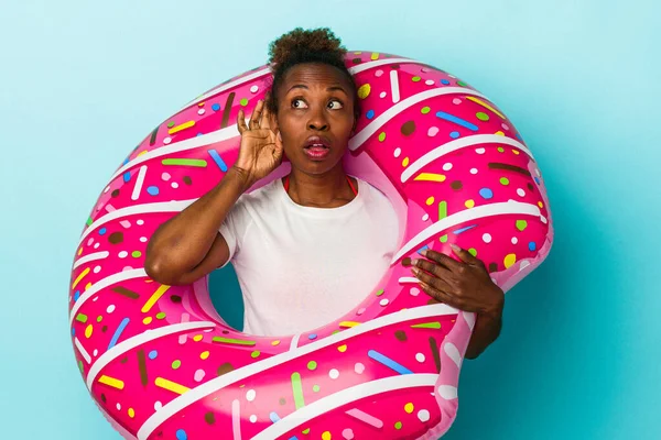 Young african american woman with inflatable donut isolated on blue background trying to listening a gossip.