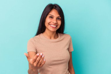 Mavi arka planda izole edilmiş genç Latin kadın selamlama jesti olarak kameraya elini uzatıyor..