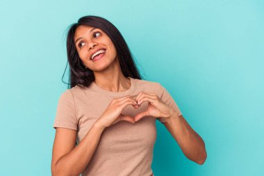 Mavi arka planda izole edilmiş genç Latin kadın gülümsüyor ve elleriyle kalp şeklini gösteriyor..