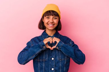 Pembe arka planda izole edilmiş genç melez kadın gülümsüyor ve elleriyle kalp şeklini gösteriyor..