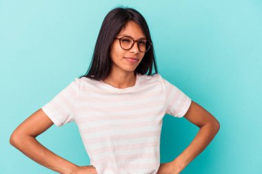 Mavi arka planda izole edilmiş, hoşnutsuzca kaşlarını çatan, kollarını kavuşturan genç Latin bir kadın..