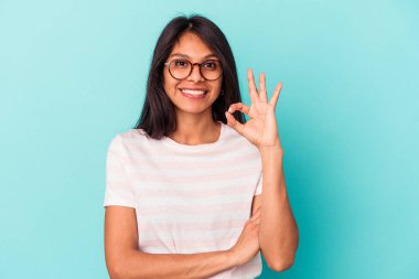Mavi arka planda izole edilmiş genç Latin bir kadın bir göz kırpıyor ve elleriyle iyi bir jest yapıyor..