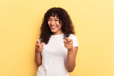 Sarı arka planda izole edilmiş genç melez kadın parmaklarıyla ön tarafı işaret ediyor..